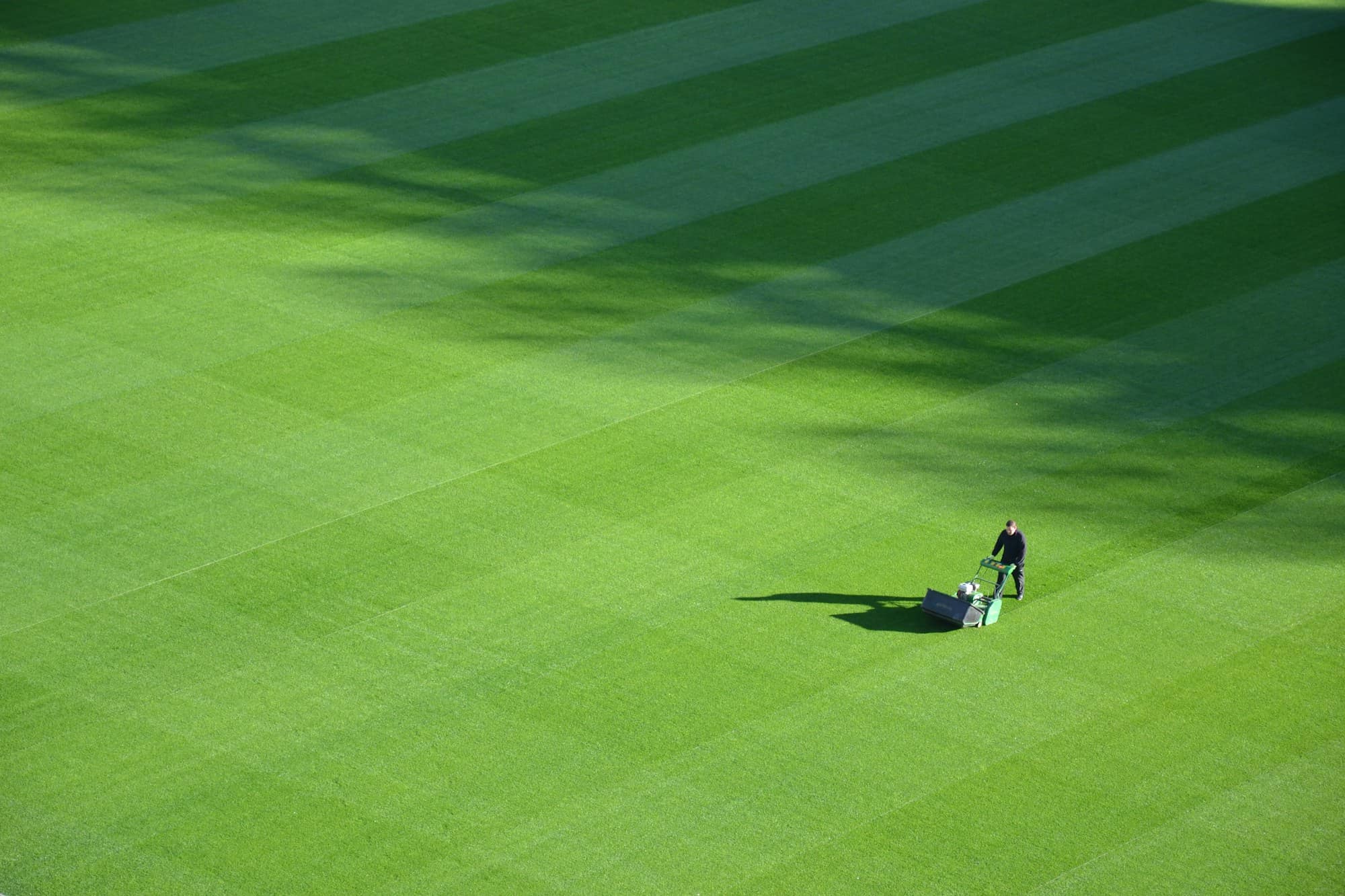 Aerial view of a large striped sports field with a single groundskeeper pushing a mower across it