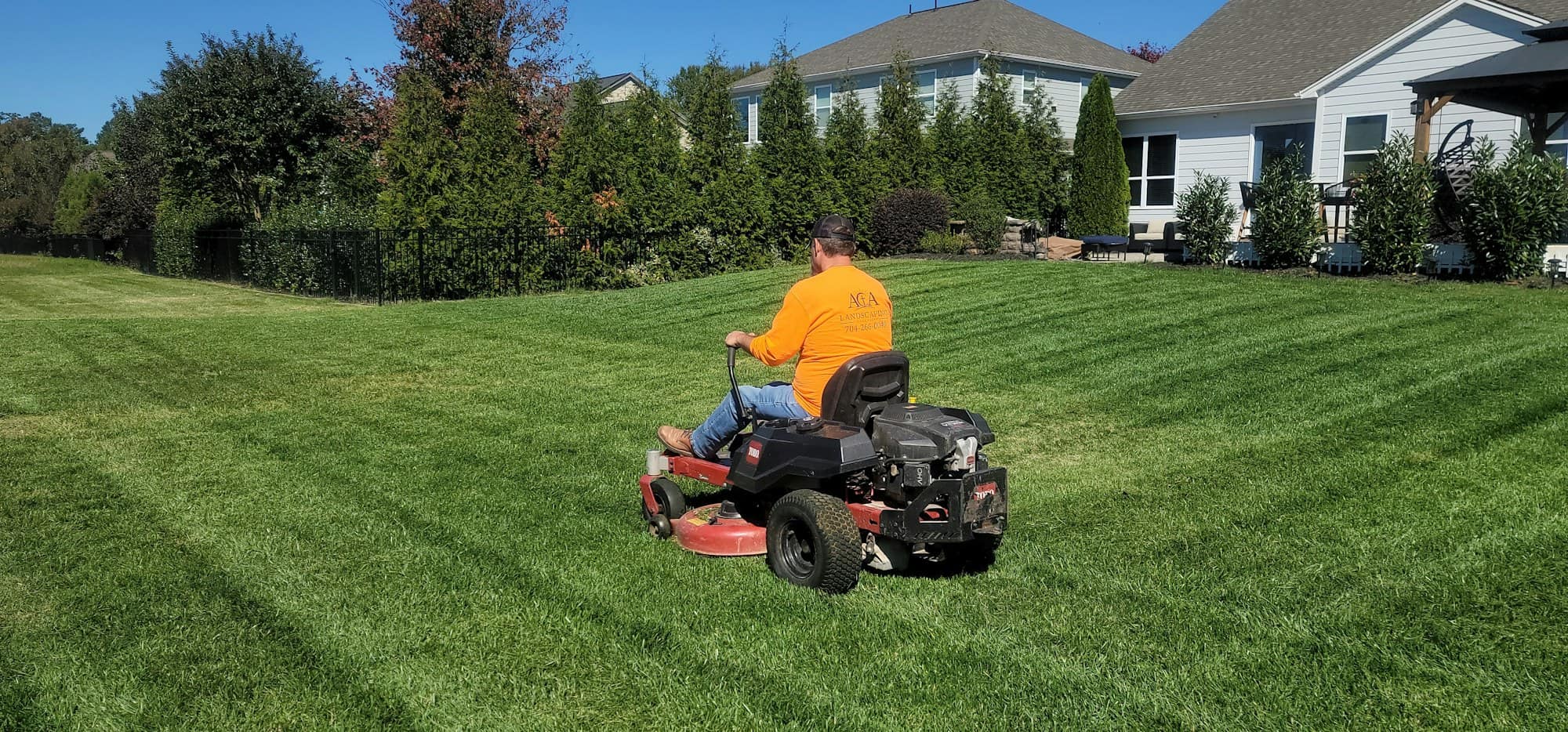 Operator on a zero-turn ride-on mower cutting stripes into a wide lawn with houses behind